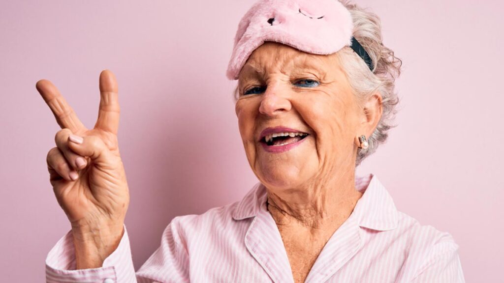 Eine alte Frau in rosa Pyjama und rosa Plüschmaske auf der Stirn zeigt ein Victory Zeichen. (c) AdobeStock