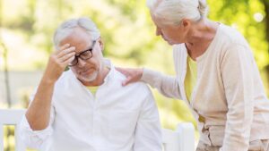 Ein Mann mit Brille auf einer Bank sitzend mit Kopfschmerzen; daneben eine Frau, die die Hand auf seine Schulter legt. (c) AdobeStock