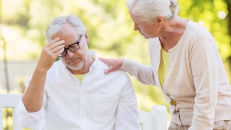 Ein Mann mit Brille auf einer Bank sitzend mit Kopfschmerzen; daneben eine Frau, die die Hand auf seine Schulter legt. (c) AdobeStock