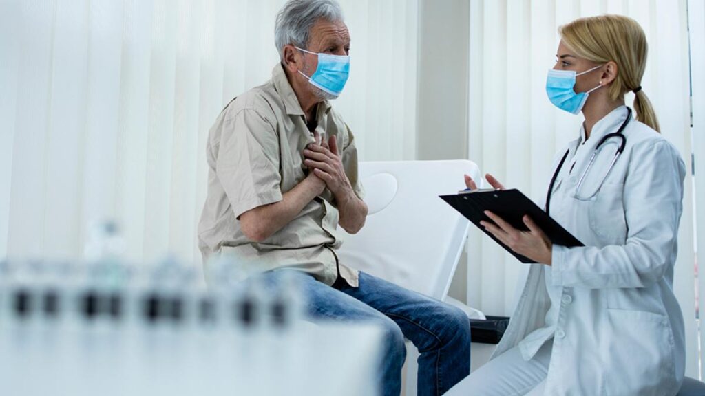 Ein Mann mit Gesichtsmaske sitzt neben einer Ärztin auf dem Behandlungsbett und greift sich mit beiden Händen an die Brust. (c) AdobeStock