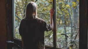 Eine Frau steht beim Fenster und schaut hinaus. (c) AdobeStock