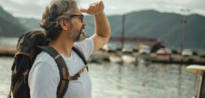 Ein Mann mit Sonnenbrille, Vollbart und Rucksack vor einem See in die Ferne schauend. (c) AdobeStock