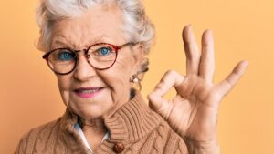 Eine ältere Frau mit Brille zeigt zufrieden lächelnd ein "alles ok"-Zeichen mit ihrer linken Hand. (c) AdobeStock